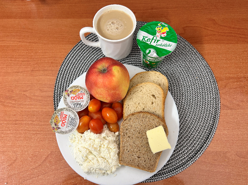 Na zdjęciu znajduje się: Kefir 2%, Kawa zbożowa z mlekiem, Chleb mieszany pszenno-żytni, Chleb Graham, Masło extra 82%, Miód, Twarożek, Jabłko, ​​​​​​​Pomidorki koktajlowe